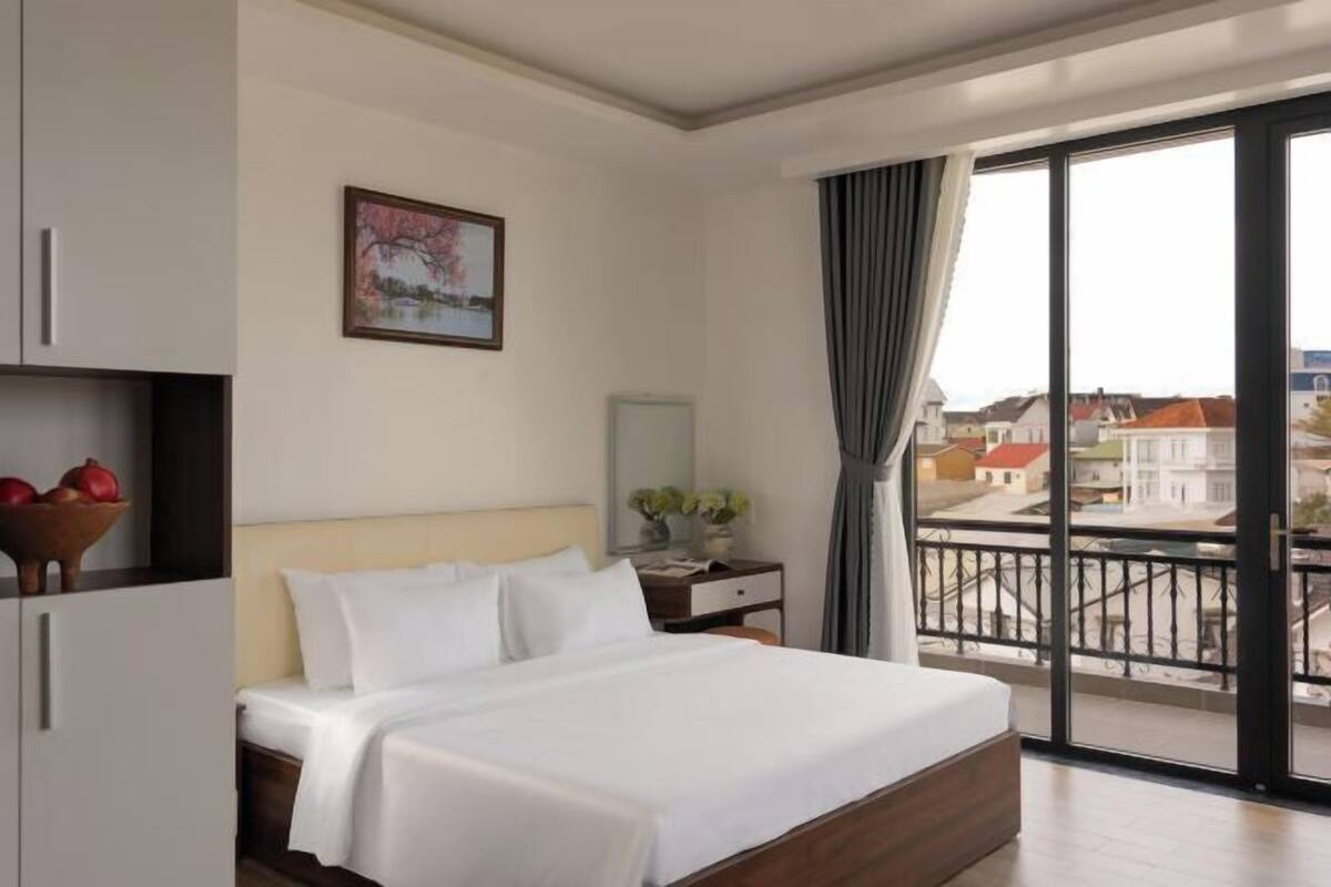 A hotel room with a bed, a nightstand, a window with a view of buildings, and a painting on the wall.