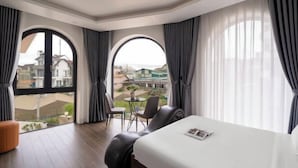 A hotel room with a large bed, two chairs, a small table, and a view of the city through large windows. - Anteaus Hotel & Apartment Da Lat (Dalat)