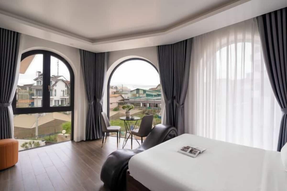 A hotel room with a large bed, two chairs, a small table, and a view of the city through large windows.