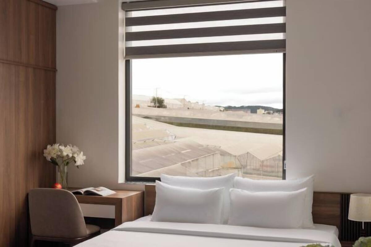 A hotel room with a large window, a bed with white linens, a desk with a chair, and a vase with flowers.