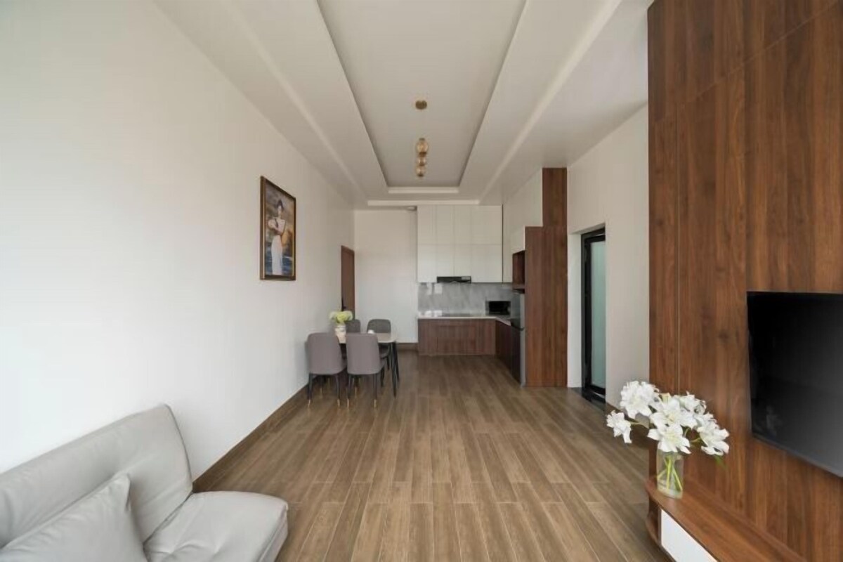 A modern living room with a sofa, a dining area with chairs, a kitchen in the background, and a flat-screen TV mounted on a wooden panel.