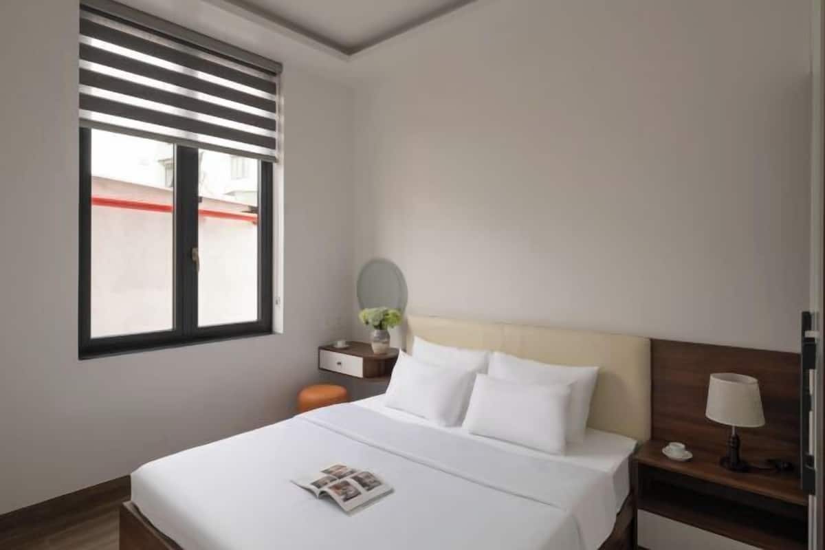 A hotel room with a bed, a bedside table with a lamp, a window with blinds, and a small round mirror.