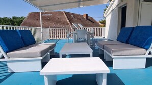 A covered outdoor seating area with white furniture and blue cushions, a table, and a view of a house with a tiled roof. - Apartments Mateo - One Bedroom Apartment - Blue (Vodice)