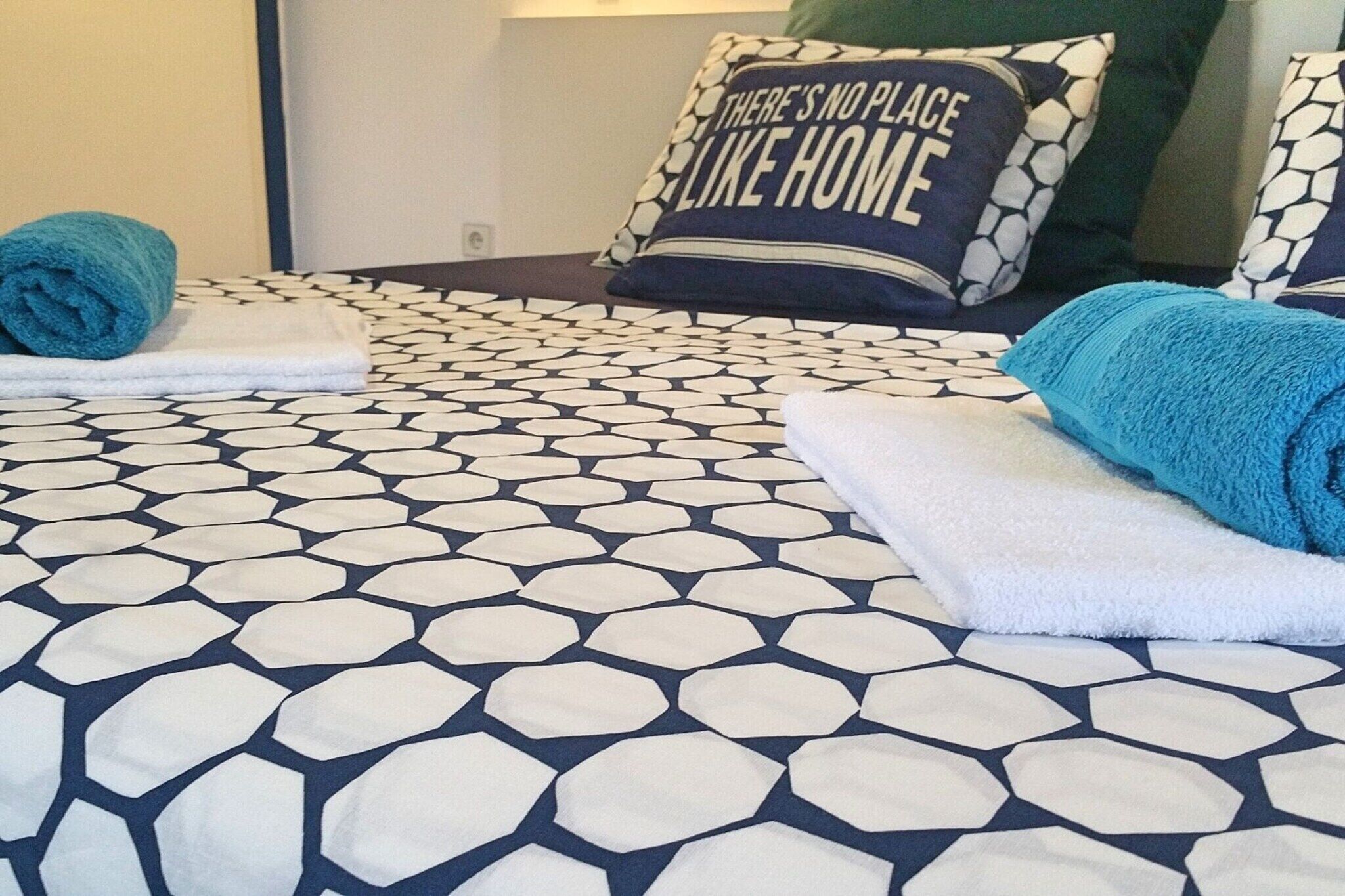 A bed with a geometric patterned bedspread, two rolled towels, and a pillow with the text "THERE'S NO PLACE LIKE HOME".