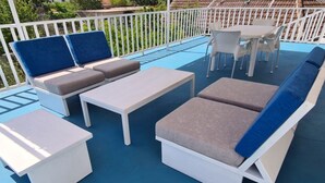 A covered outdoor seating area with white furniture and blue cushions, a table, and a white fence. - Apartments Mateo - One Bedroom Apartment - Blue (Vodice)