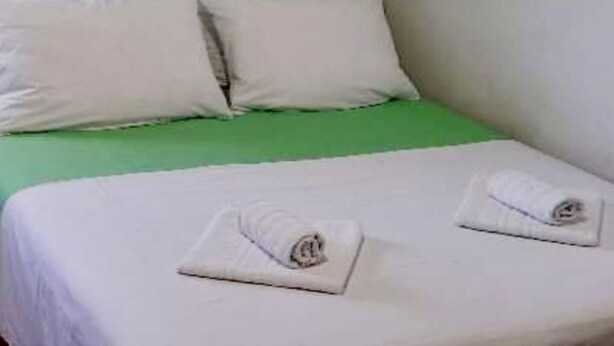 A single bed with white bedding and pillows, a green headboard, and two rolled towels on the bed.
