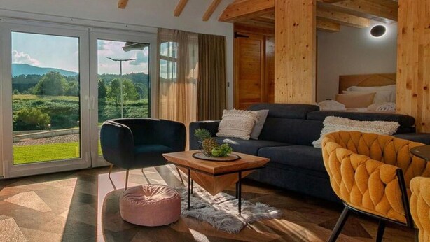 Una sala de estar moderna con un sofá azul oscuro, una mesa de centro de madera y un sillón amarillo. Amplias ventanas ofrecen vistas a un paisaje verde.