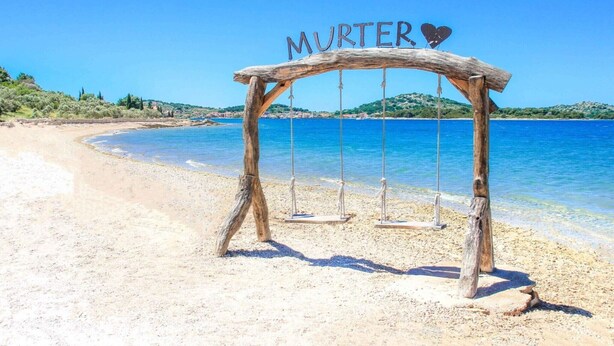 Eine Holzschaukel mit der Aufschrift "MURTER" und einem Herzsymbol, auf einem Sandstrand mit klarem, blauen Wasser und Hügeln im Hintergrund.