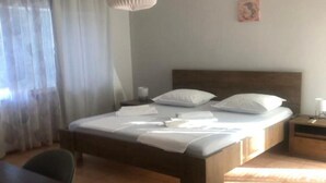 A neatly made bed with pillows and a blanket, a bedside table with a lamp, and a chair in a room with a tiled floor and a window with curtains.