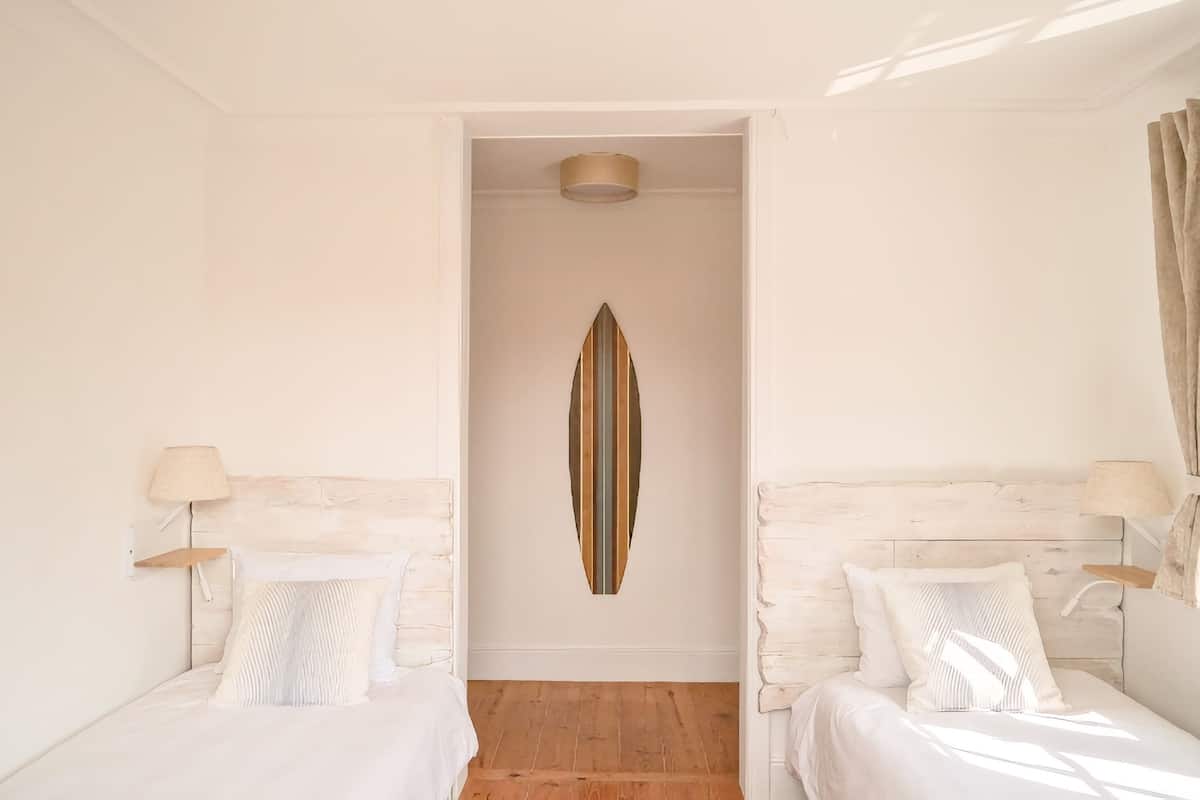 A room with two beds, a wooden floor, a wall-mounted surfboard decoration, and a ceiling light.