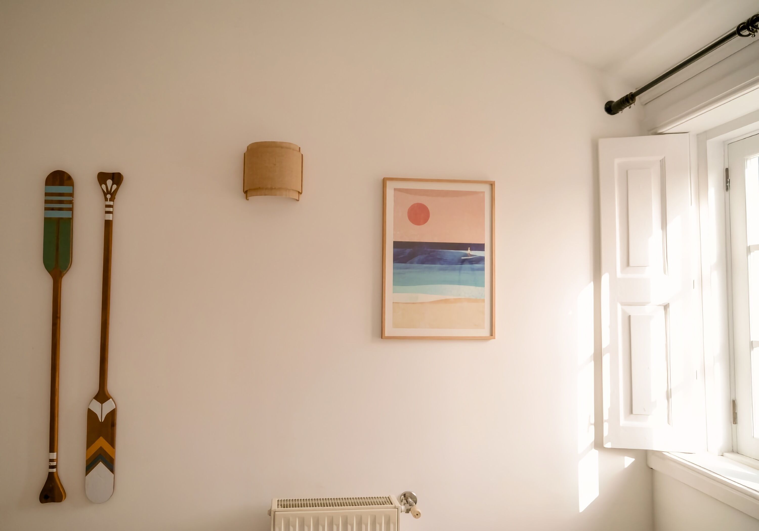 A room with a white radiator, a framed picture of a beach scene, and two wooden paddles on the wall.