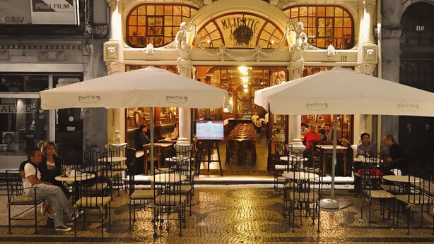 An ornate building with a marquee sign reading "MAJESTIC" and a number "112" above the entrance. There are outdoor tables and chairs under white umbrellas, and people are seated and walking around. - Porto Surf Lodge (Porto)