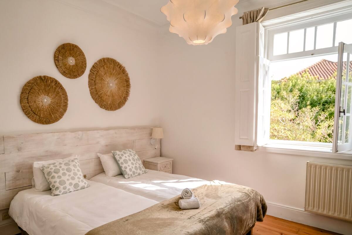 A bedroom with a large bed, a nightstand, a wall-mounted lamp, and a view of greenery outside.