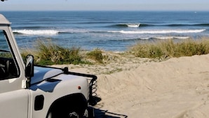 A white vehicle parked on a sandy beach with ocean waves in the background. - Porto Surf Lodge (Porto)