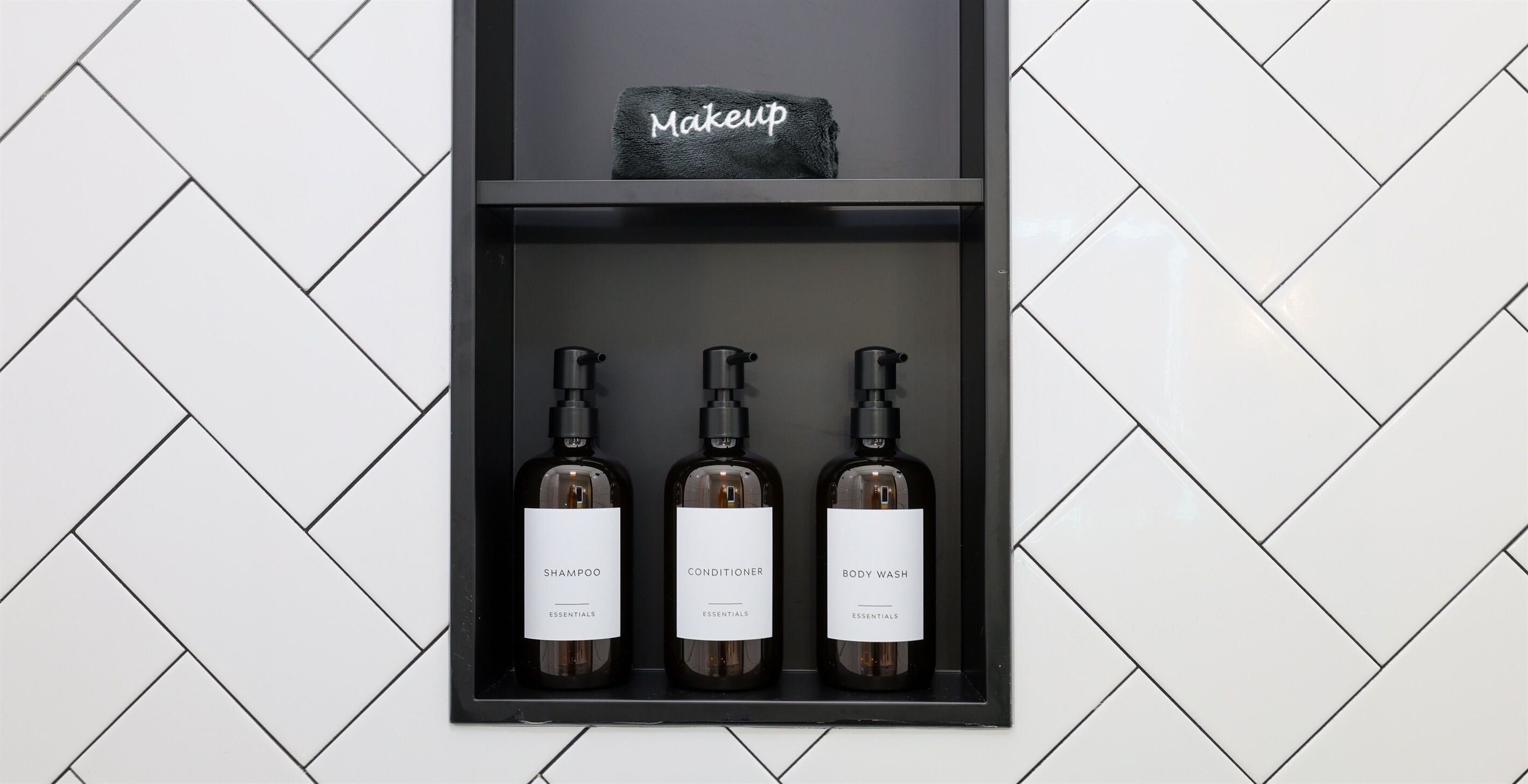 Ein Regal mit drei Flaschen mit den Aufschriften 'SHAMPOO', 'CONDITIONER' und 'BODY WASH' von 'ESSENTIALS'. Darauf ein schwarzes Handtuch mit dem Schriftzug 'Makeup'.