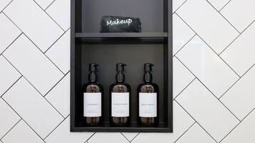 Ein Regal mit drei Flaschen mit den Aufschriften 'SHAMPOO', 'CONDITIONER' und 'BODY WASH' von 'ESSENTIALS'. Darauf ein schwarzes Handtuch mit dem Schriftzug 'Makeup'.
