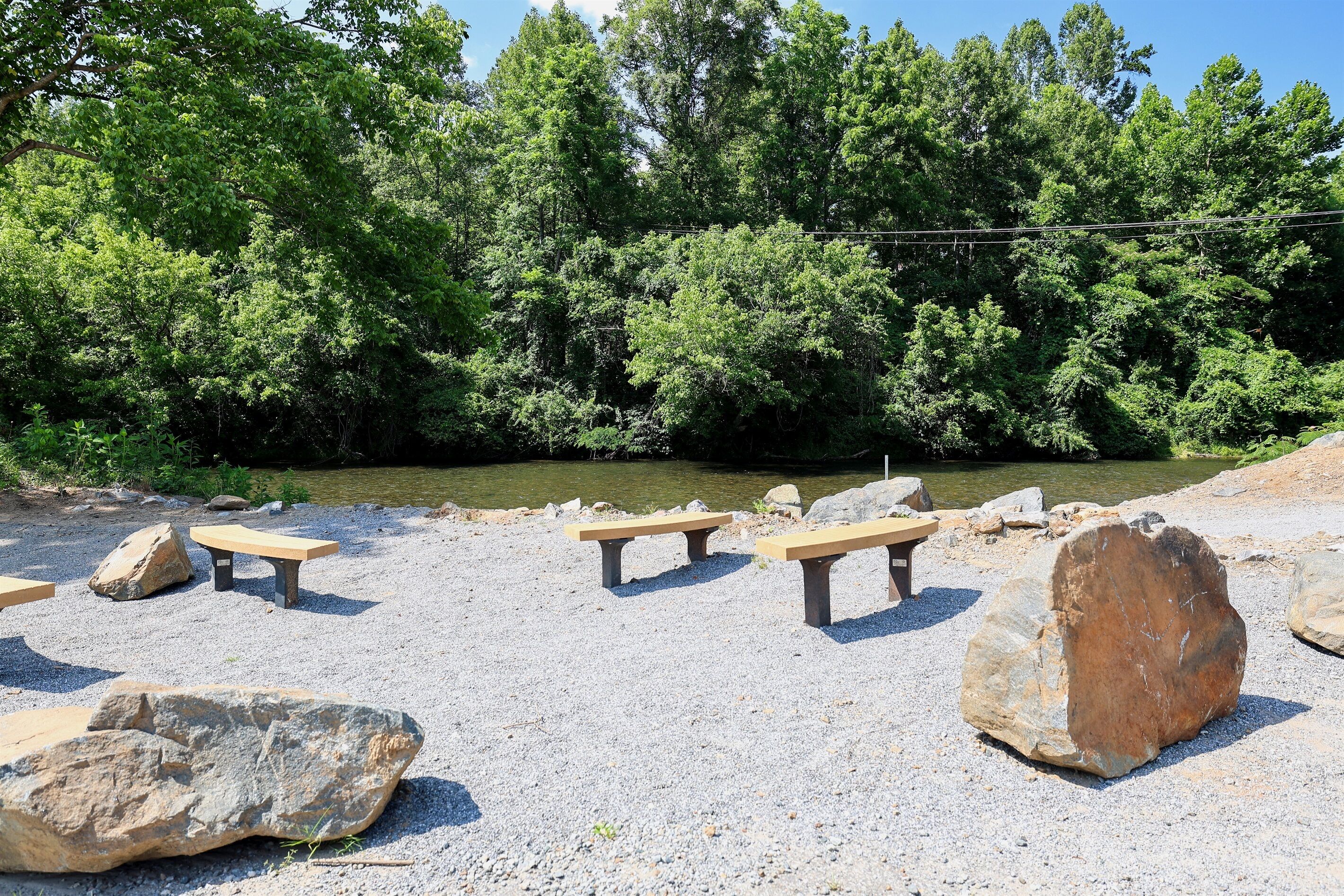 A natural setting with picnic tables and large rocks, surrounded by dense green trees and a calm water body.