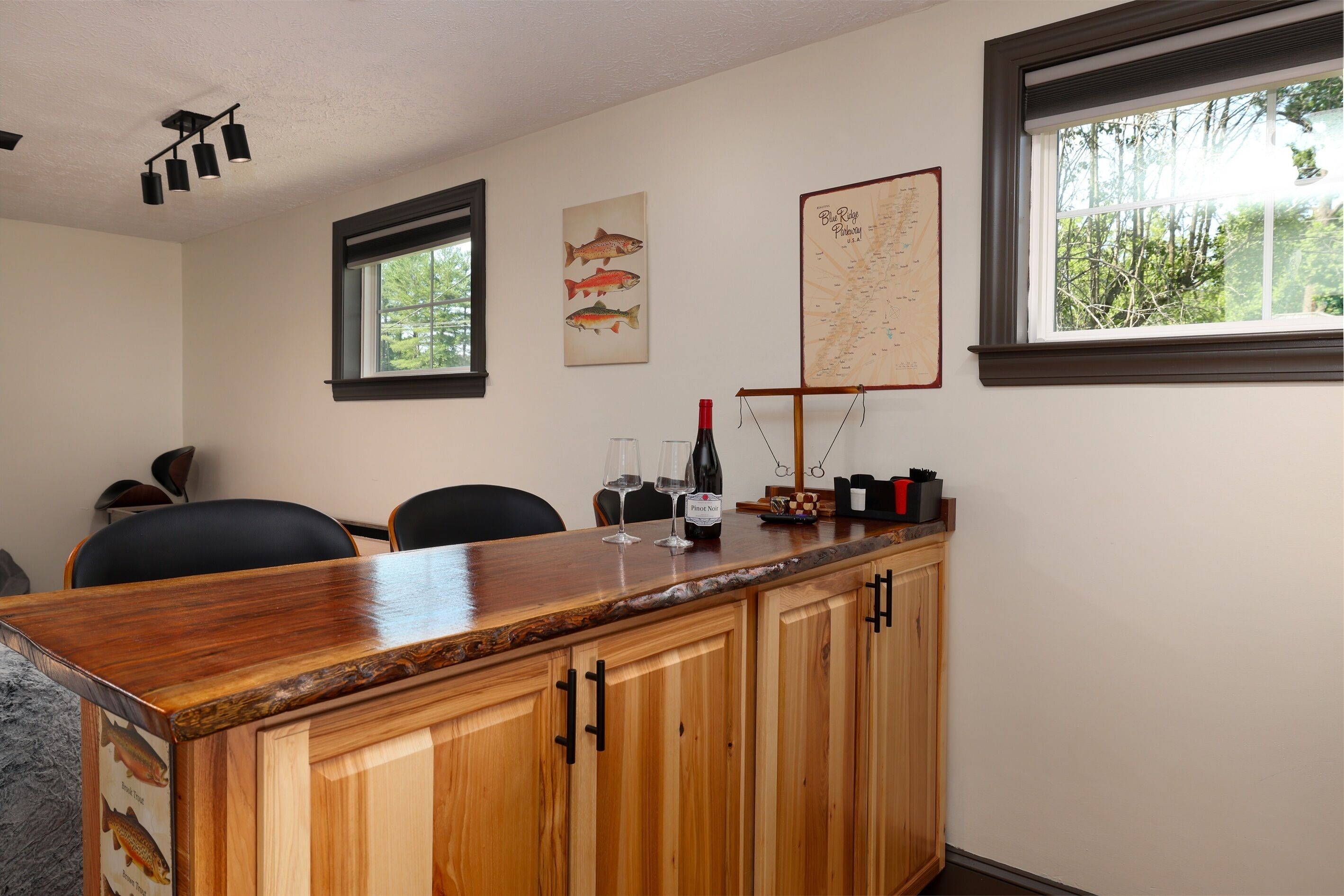 Una zona de bar con barra de madera, un mapa e ilustraciones de peces en la pared, una ventana con vista a árboles y una botella de vino.