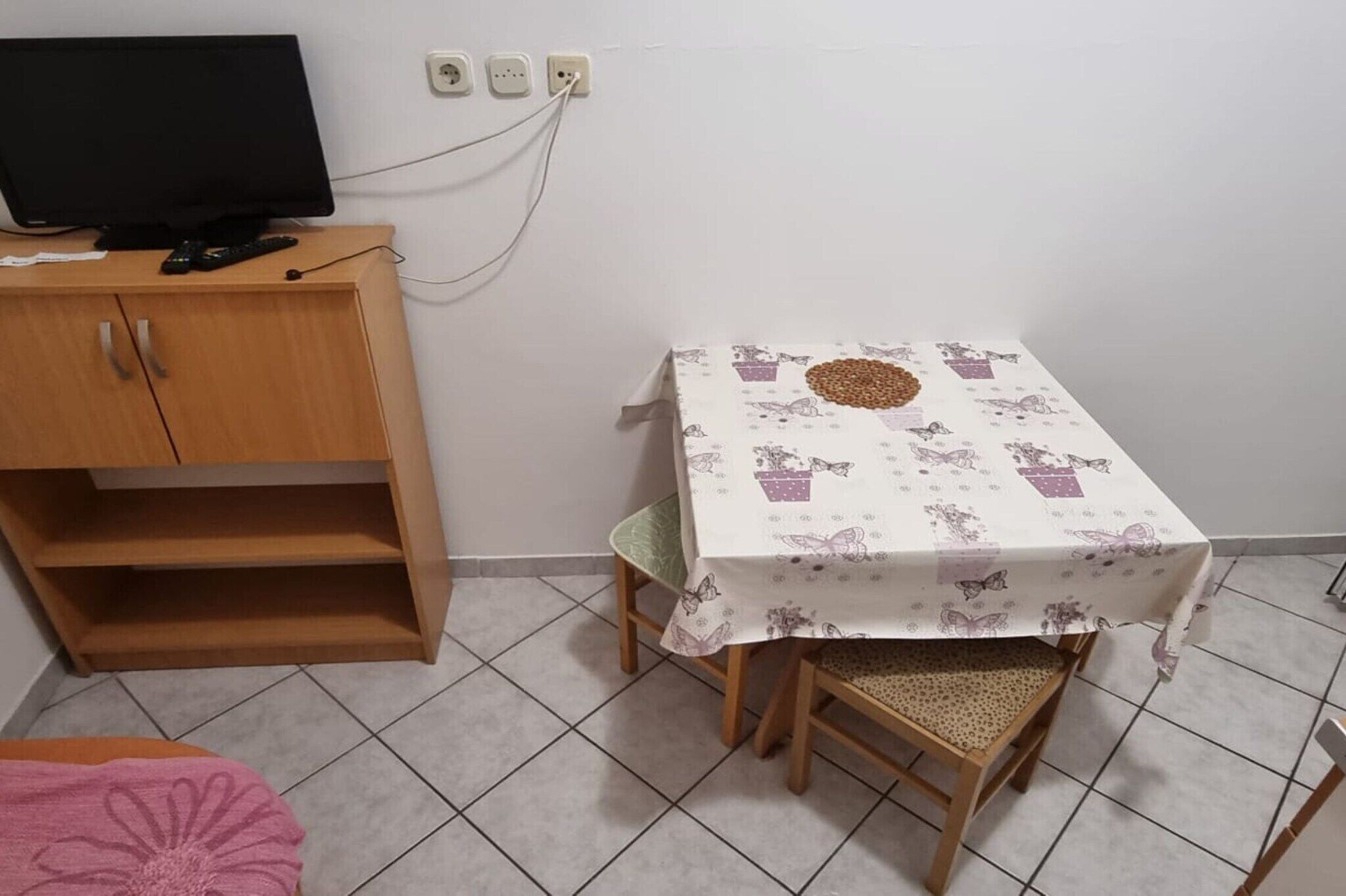 Een kleine kamer met een houten kastje, een televisie erop, een tafel met een bloemenkleed en een stoel.