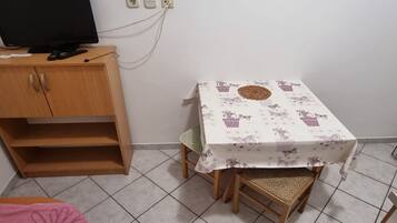 Een kleine kamer met een houten kastje, een televisie erop, een tafel met een bloemenkleed en een stoel.