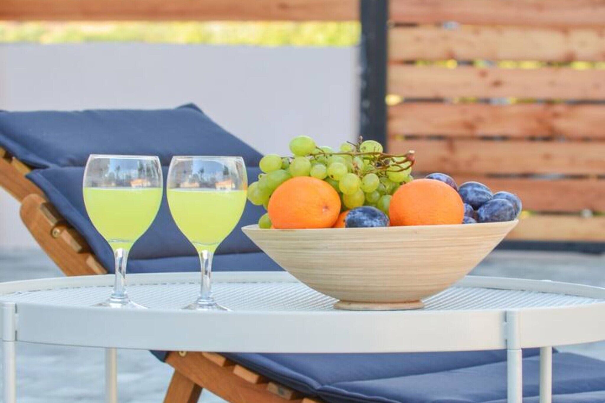 A table with two glasses of yellow drink, a bowl of fruit, and a wooden chair with a blue cushion.