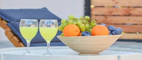 A table with two glasses of yellow drink, a bowl of fruit, and a wooden chair with a blue cushion.