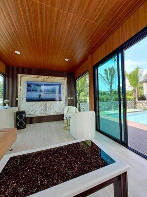 A modern living room with a large TV, comfortable seating, and a view of a swimming pool. - Naisuan villa (Trang)