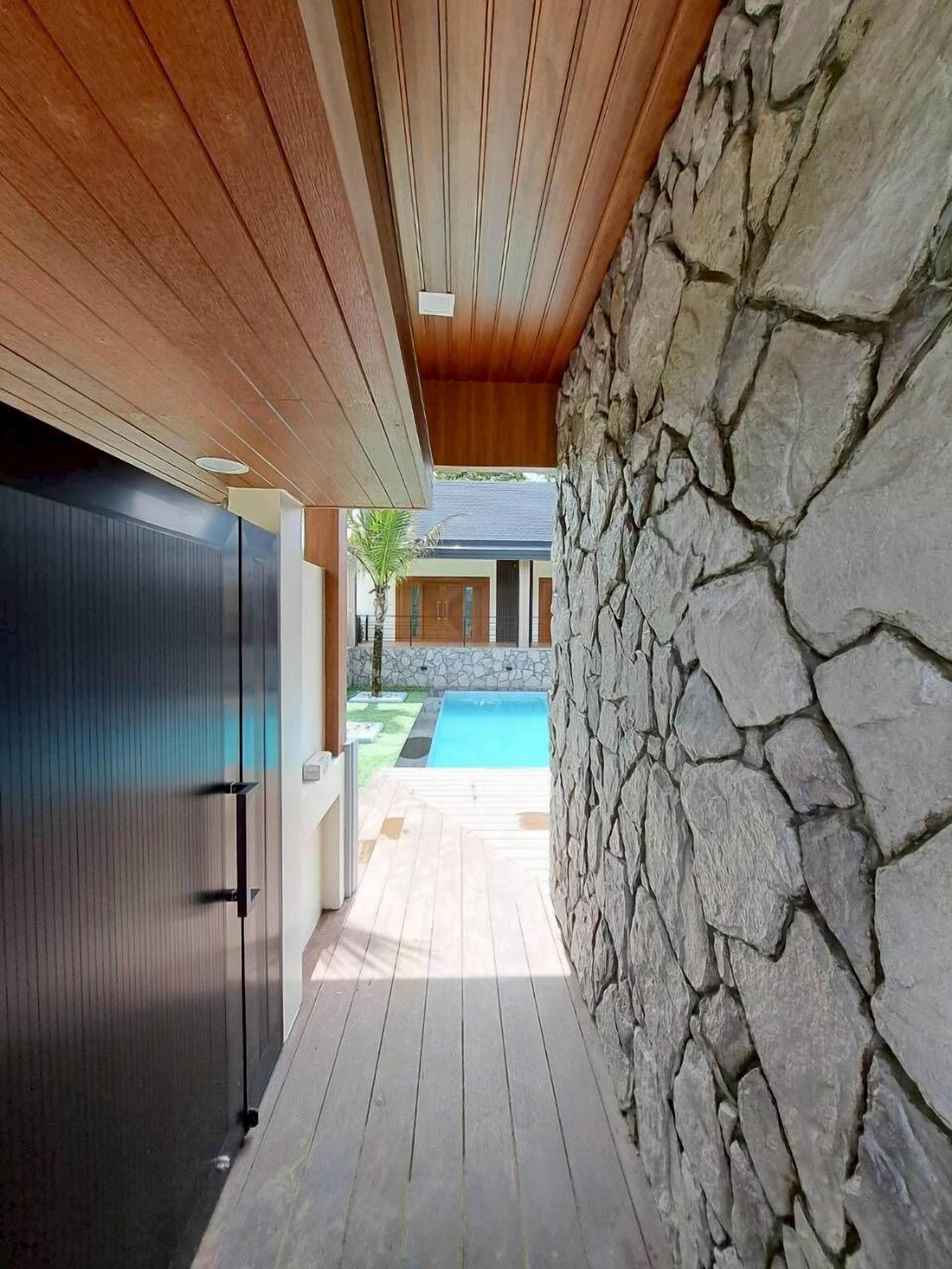 A modern outdoor corridor with a wooden ceiling, a stone wall, and a view of a swimming pool.