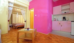 Une pièce comprenant un lit, une table à plateau en verre, un mur rose et un coin cuisine avec des placards et un évier.