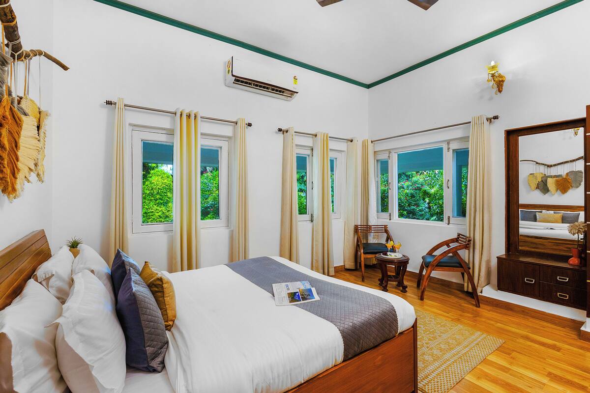 A well-lit bedroom with a large bed, a wooden headboard, a bedside table, a chair, a dresser, and a window with curtains.