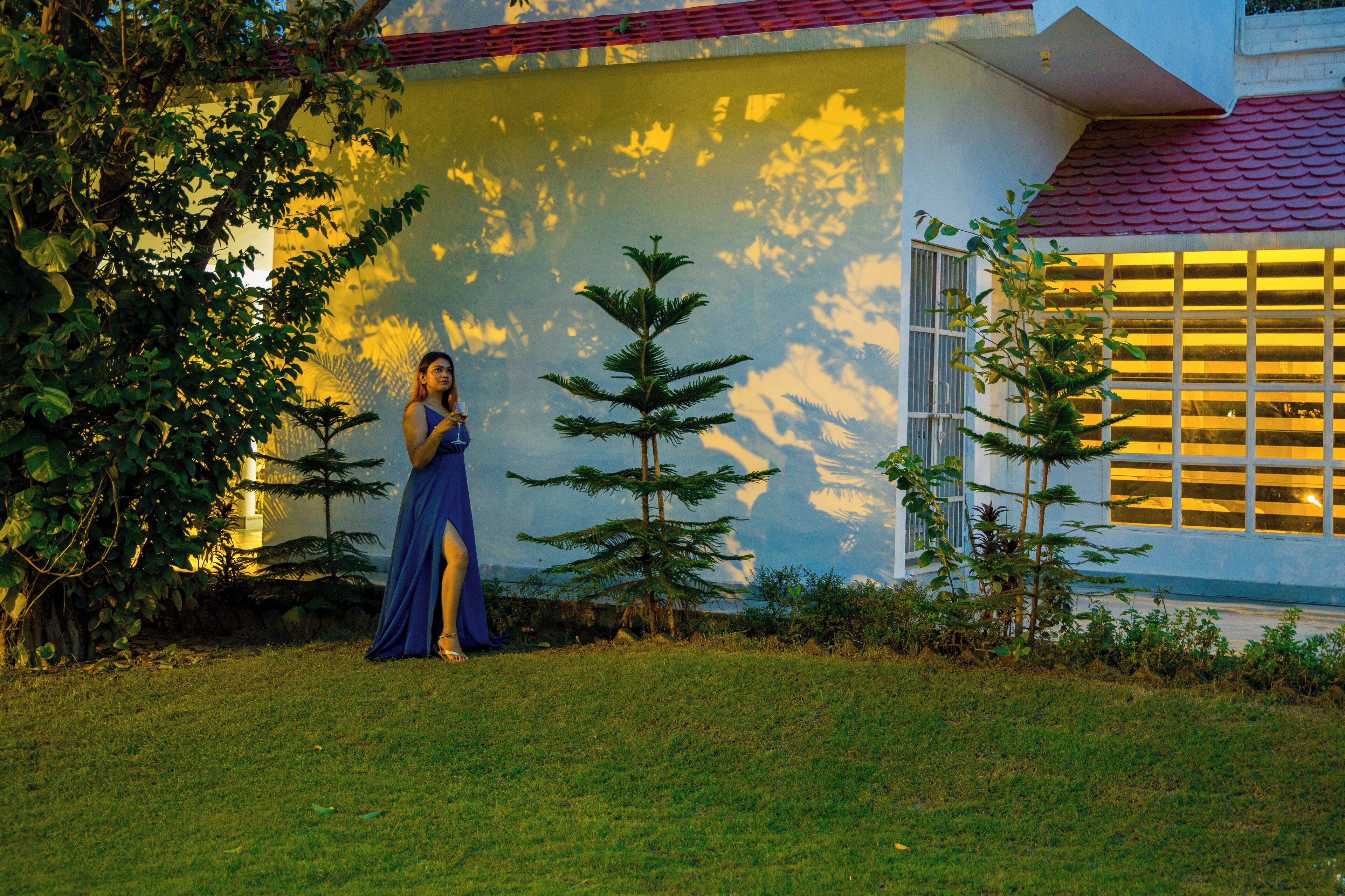 A person in a blue dress standing in a grassy area with trees and a building in the background.