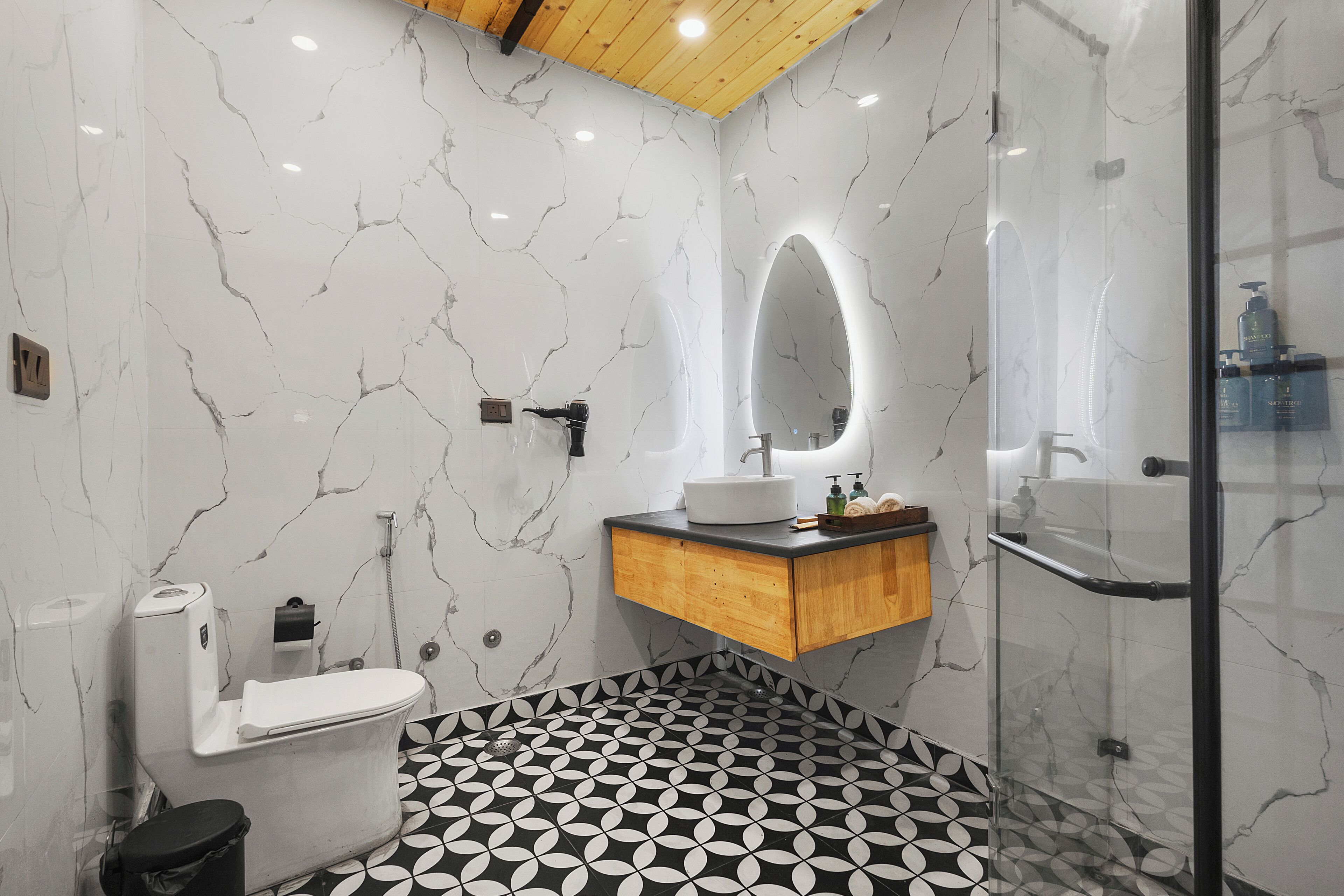 A bathroom with a marble wall, a toilet, a sink with a wooden vanity, a glass shower enclosure, and a patterned floor.