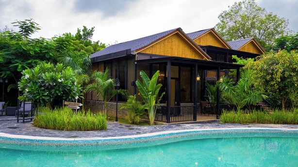 Un espace aménagé au bord de la piscine, avec un bâtiment en bois, une végétation luxuriante et une piscine à l’eau d’un bleu limpide.