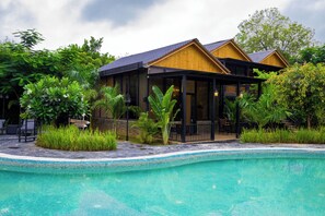 Un área junto a la piscina con un edificio de madera, abundante vegetación y una piscina azul cristalina.