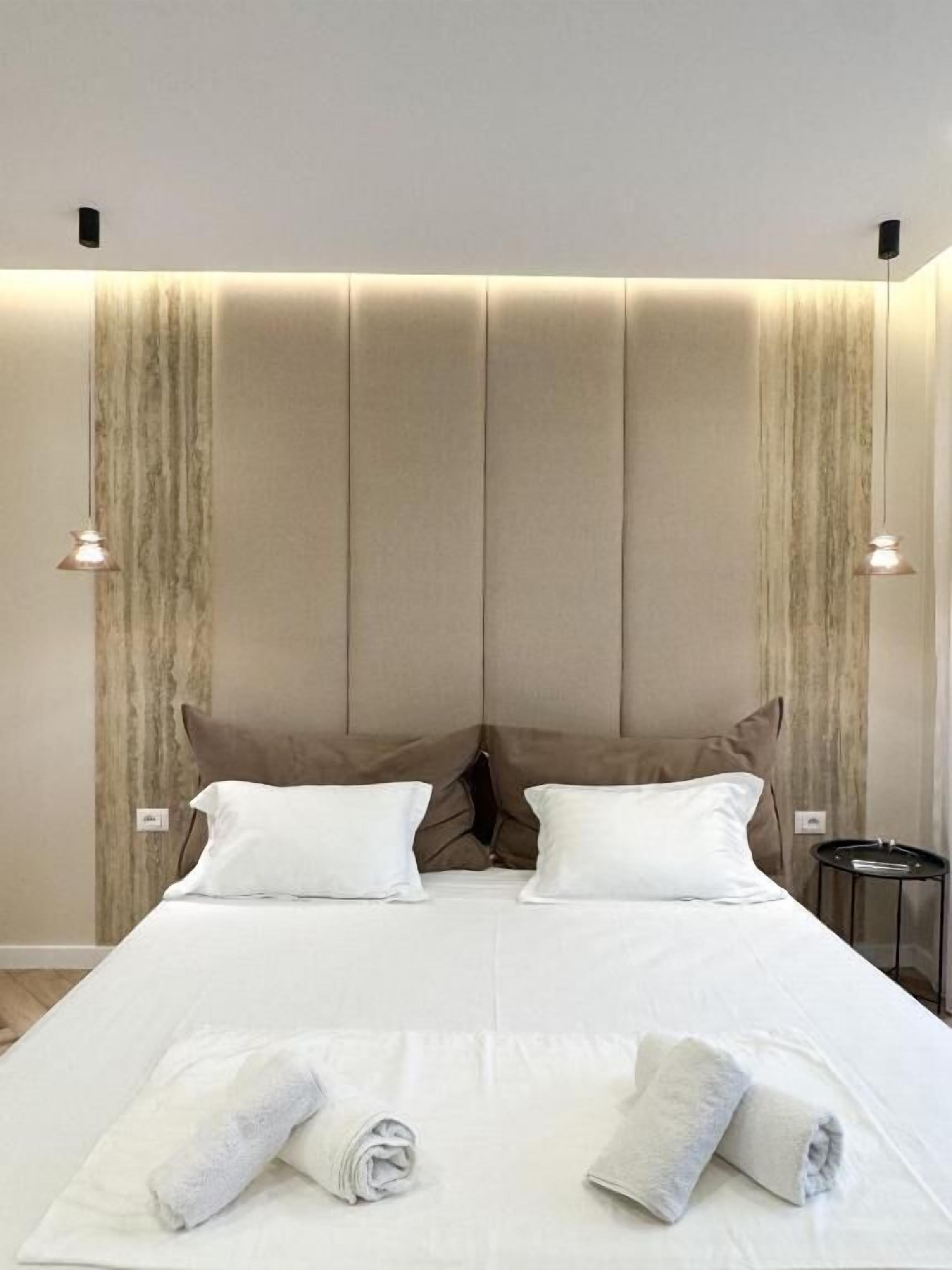 A neatly made bed with white linens and two rolled towels, a headboard with vertical panels, and two wall-mounted lamps.