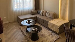 A modern living room with a sectional sofa, a round coffee table, and a large window with curtains. - Downtown Shkodra Apartment (Shkodër)