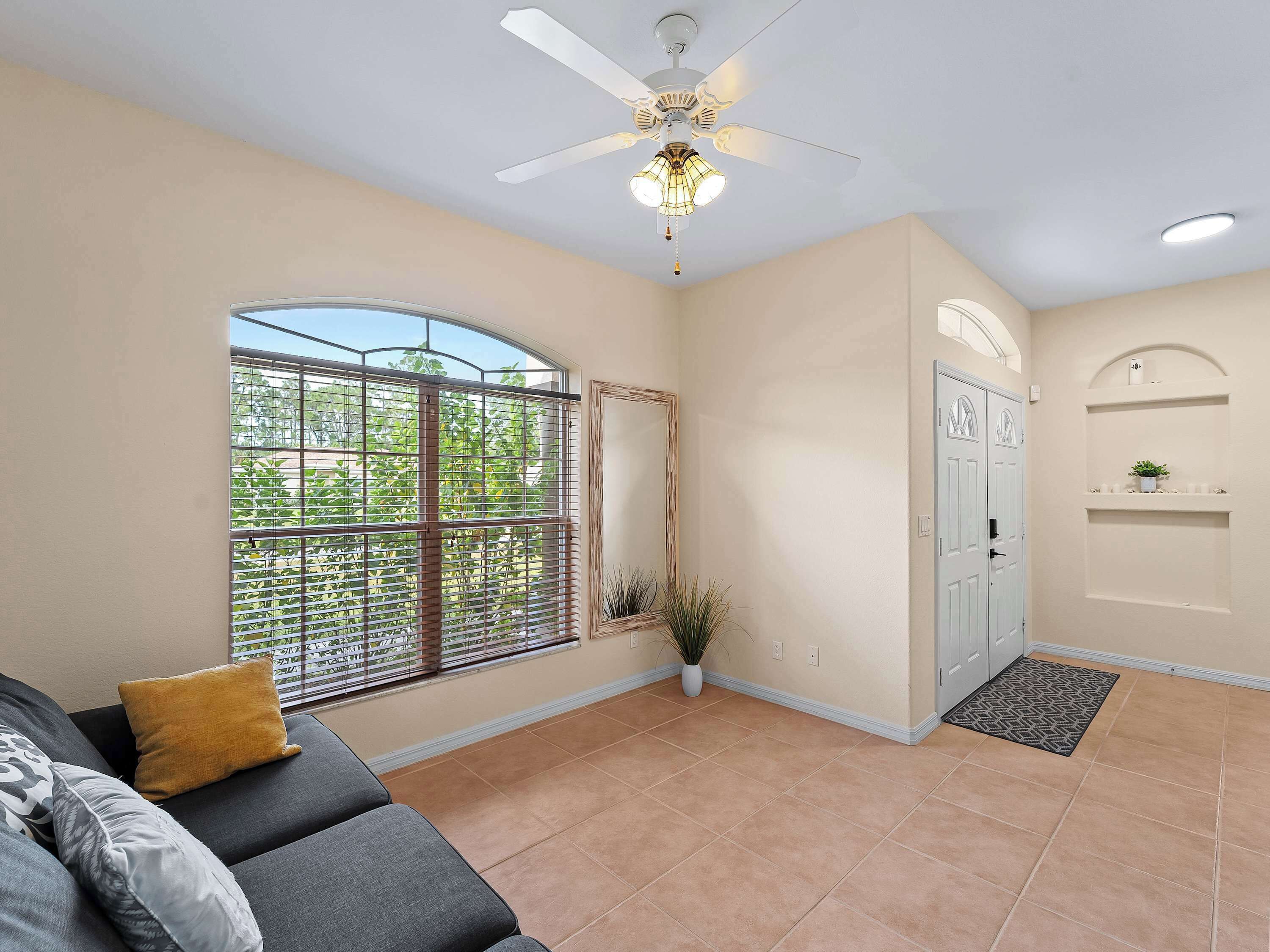 A room with a ceiling fan, a large window with a view of greenery, a door with a small shelf, and a tiled floor.