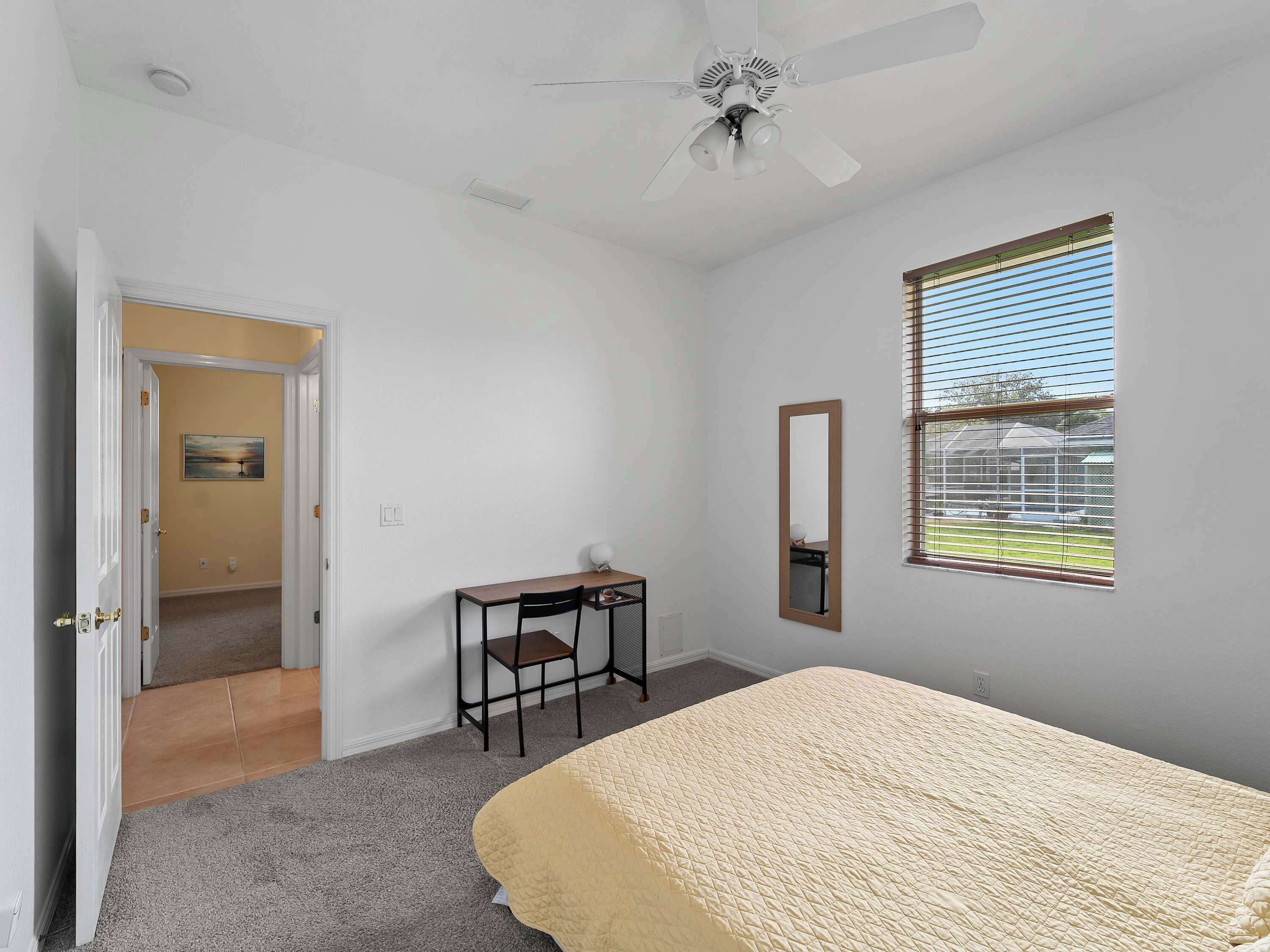 A bedroom with a bed, a desk, a chair, a ceiling fan, a window with blinds, and a door leading to another room.