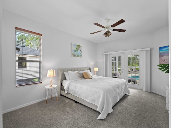 A bedroom with a bed, two bedside lamps, a ceiling fan, a window with blinds, and a door leading to a balcony.