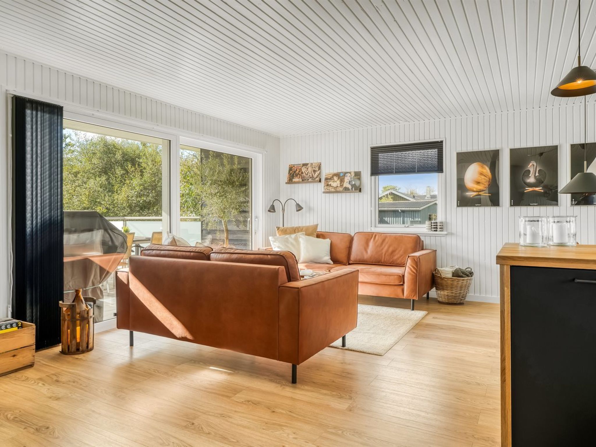 A modern living room with a brown leather sofa, a wooden coffee table, and a black cabinet. There are large windows providing natural light and a view of greenery outside.