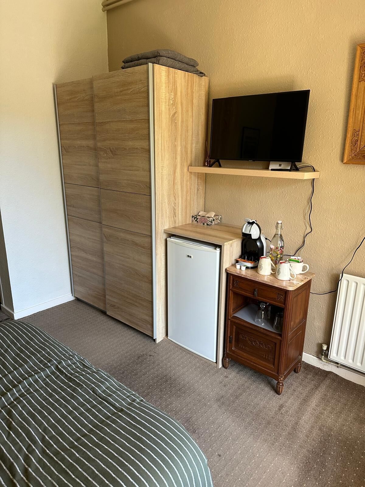 Habitación de hotel pequeña con cama, kitchenette, televisor montado en un estante de madera y radiador.