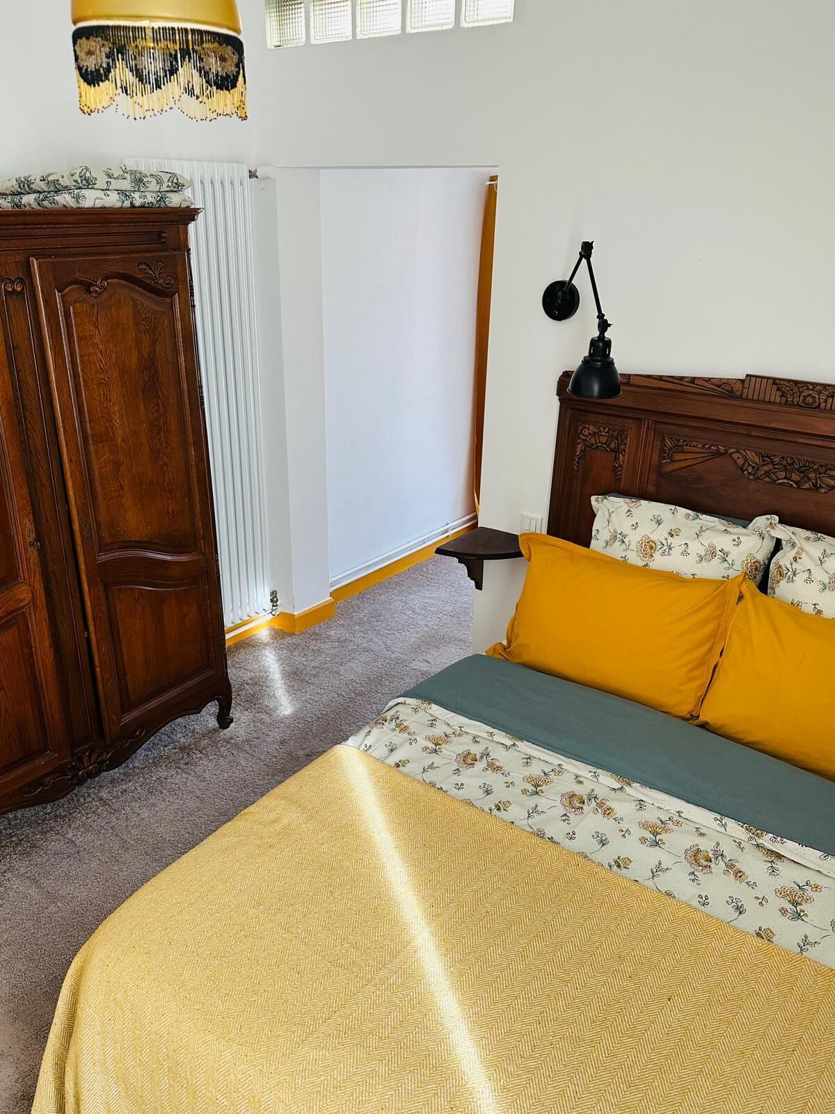 A bedroom with a wooden wardrobe, a bed with floral bedding, a bedside table with a lamp, and a whiteboard on the wall.