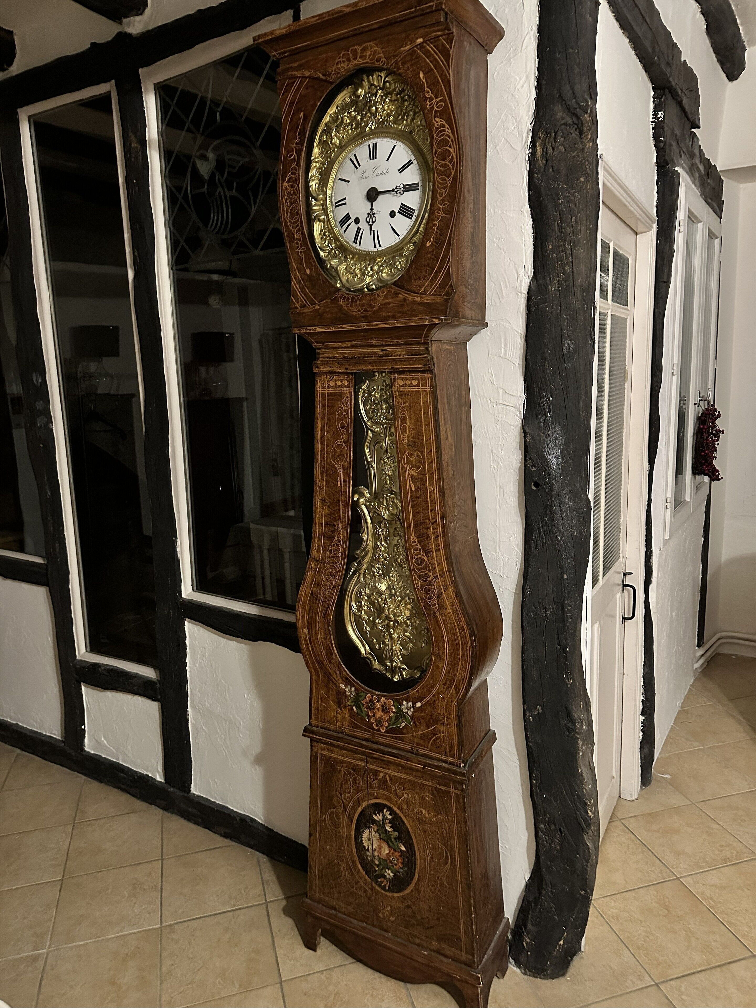 Une grande horloge grand-père, dotée de sculptures complexes et d’un grand cadran, est placée contre un mur où se trouvent une fenêtre et une porte.