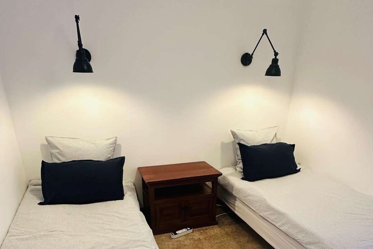 A room with two single beds, each with a pillow and a bedside table. There are two wall-mounted lamps.
