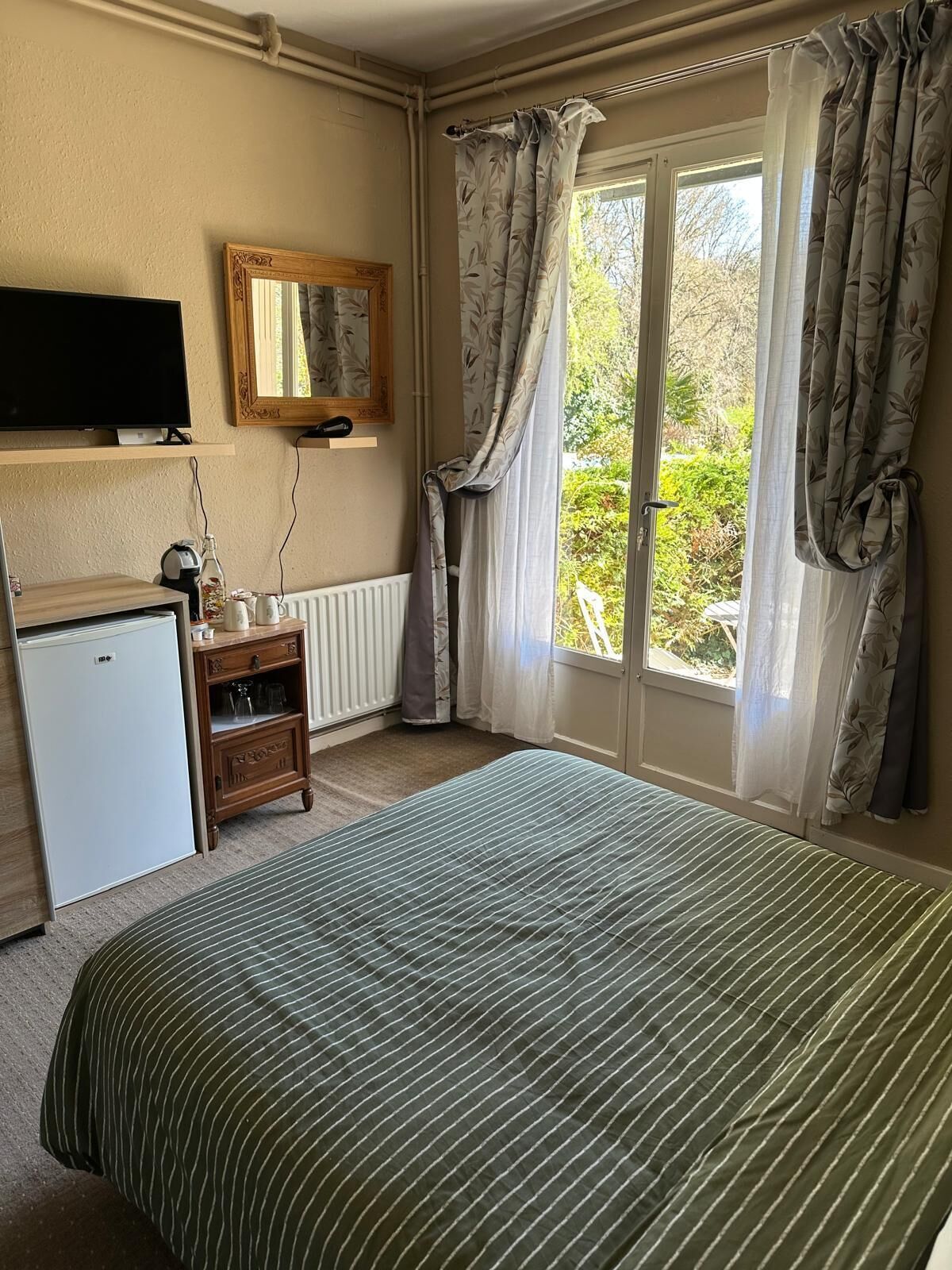 Un dormitorio con una cama, televisor, refrigerador pequeño, armario de madera y ventana con cortinas.