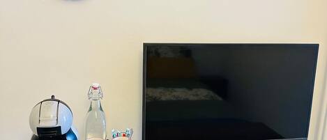 A coffee maker, a bottle, a cup, and a remote control on a wooden table with a TV in the background.