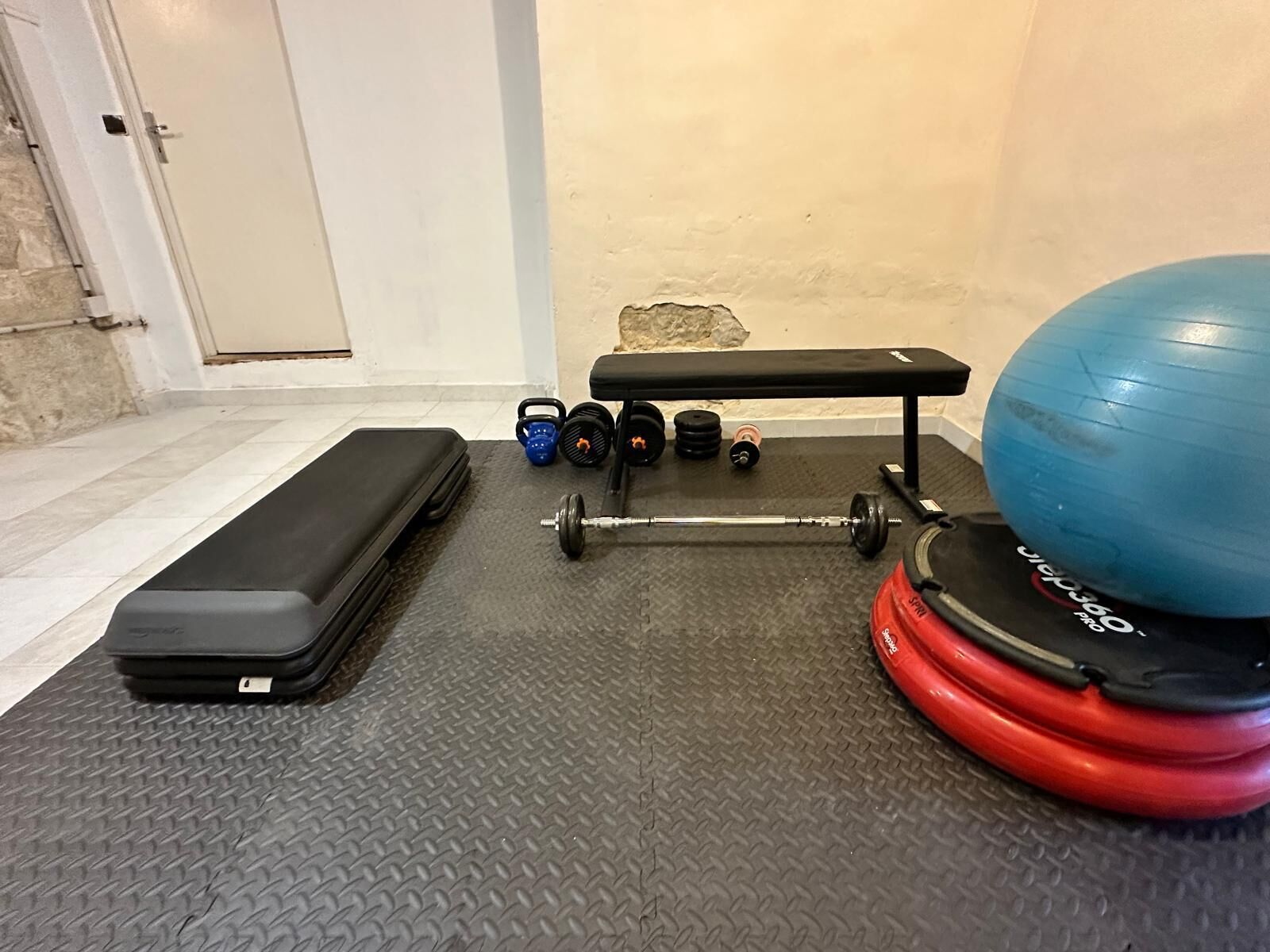 Een sportschool thuis met een bank, kettlebells, een halter, een medicine ball en een koffer.