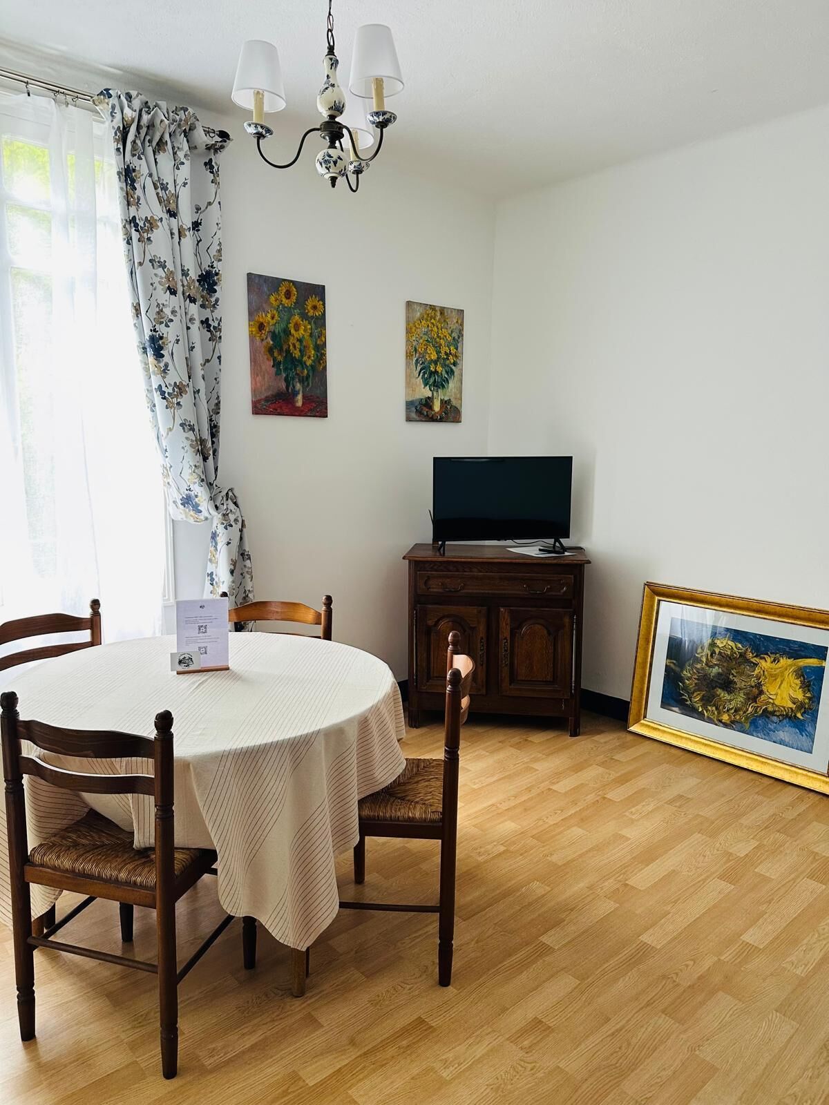 A dining area with a round table, chairs, a chandelier, paintings, a television, and a cabinet.