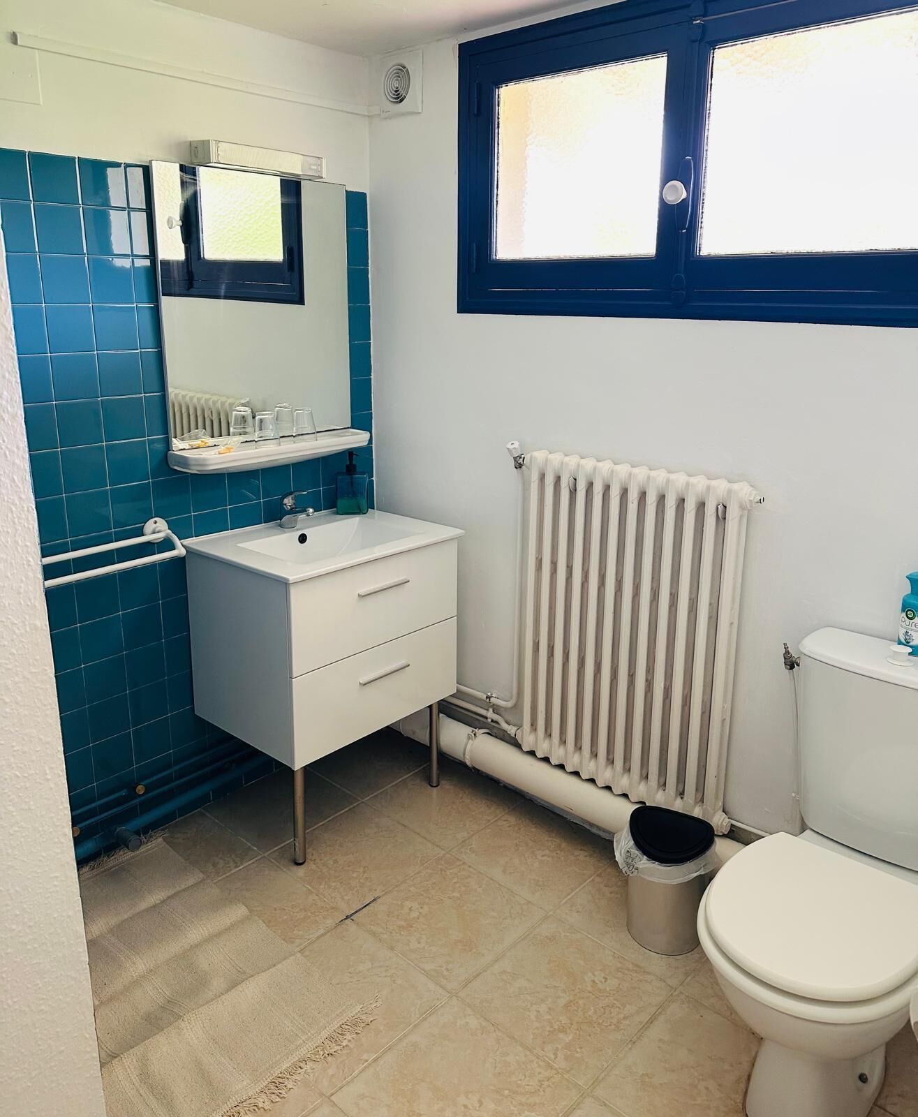 Une salle de bain avec un lavabo blanc, des carreaux bleus, un radiateur blanc et une fenêtre avec des bordures bleues.