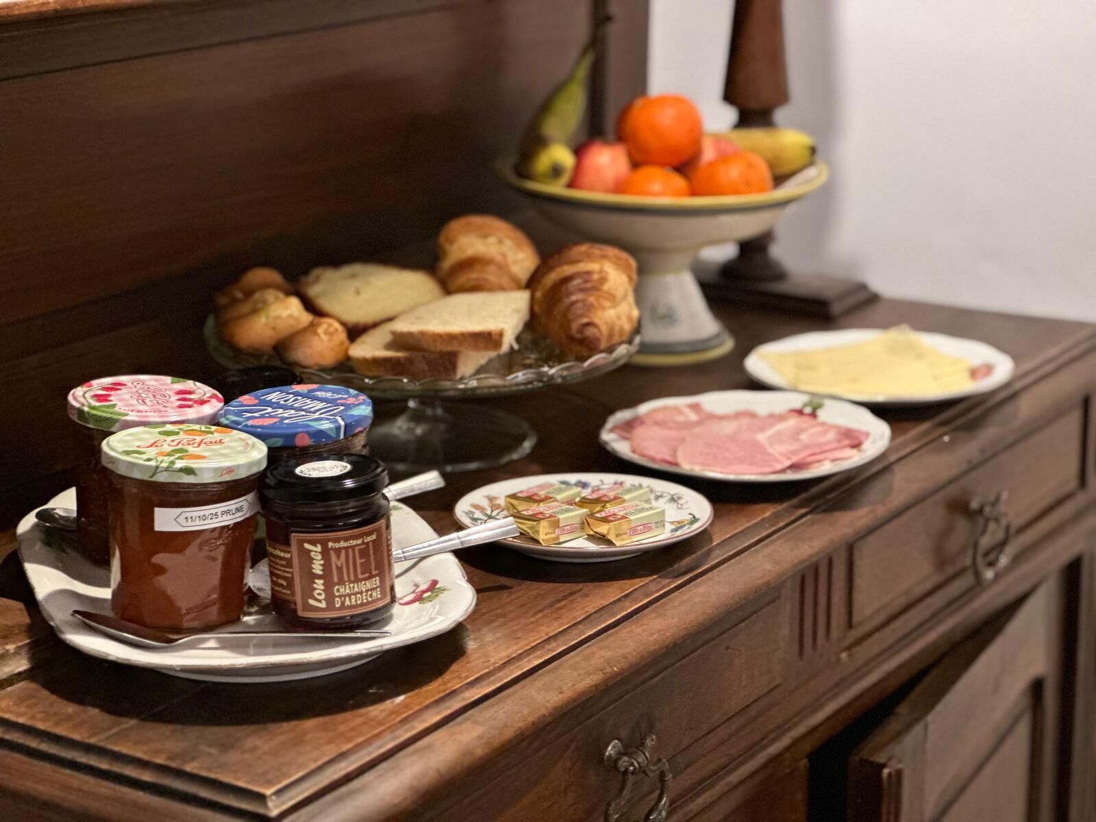 A breakfast buffet with pastries, bread, fruits, jams, cheese, and ham.